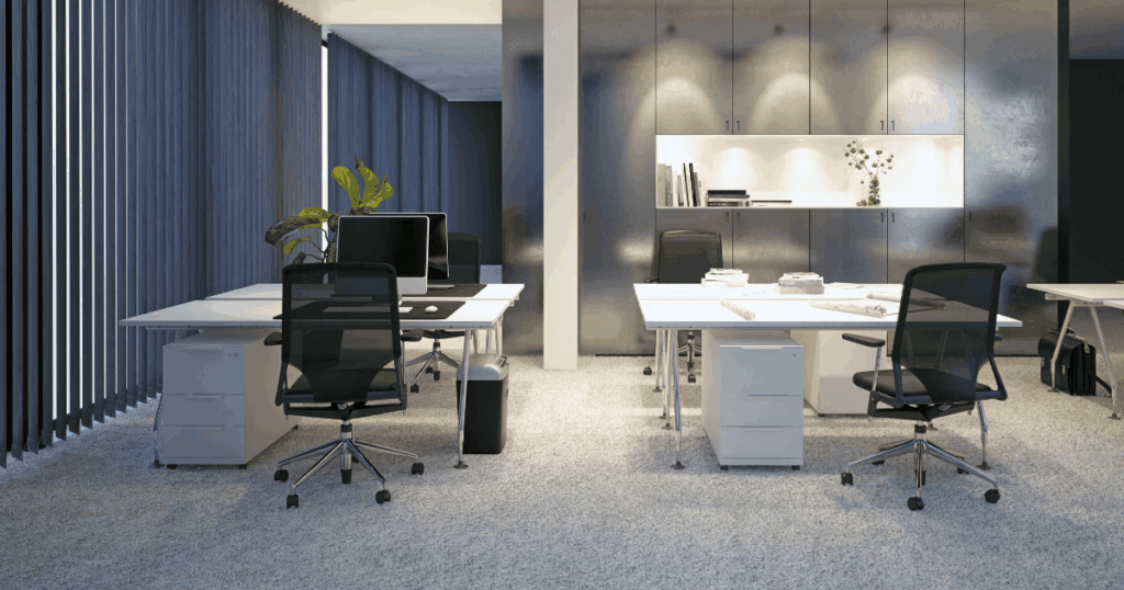 Modern open office space with ergonomic chairs, large desks, desktop computers, and natural lighting through tall vertical windows.