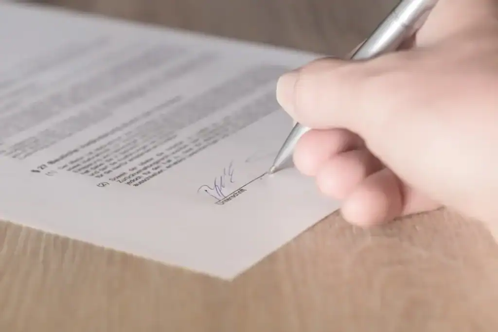 Close-up of a hand signing a printed document with a silver pen on a wooden desk.