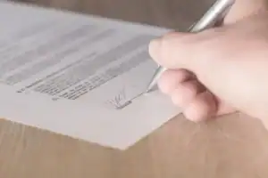 Close-up of a hand signing a printed document with a silver pen on a wooden desk.