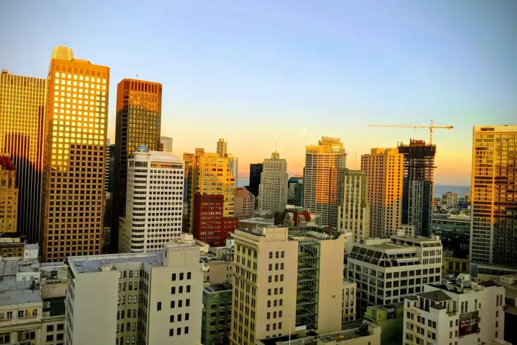 Downtown city skyline at sunset with tall office buildings glowing in golden light, construction crane visible on the right, and a clear sky overhead.