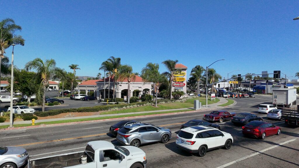 Wide street view of a KEYZ Commercial property listing featuring a Spanish style retail center with palm trees, heavy traffic, and strong street visibility near a busy intersection.